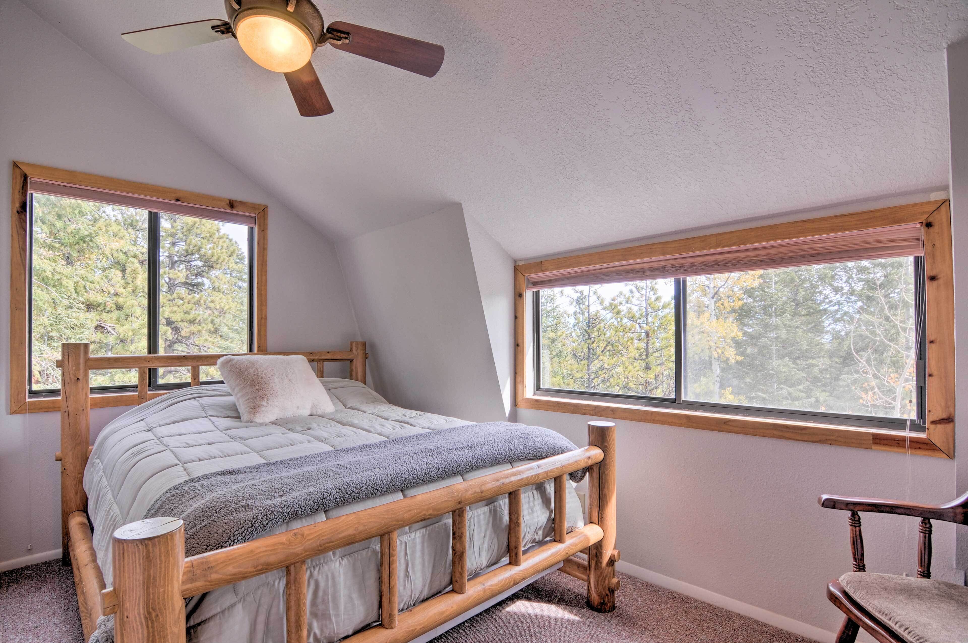 Une chambre avec un lit en bois, deux fenêtres, un ventilateur de plafond et une vue sur les arbres.