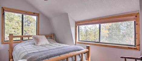 Une chambre avec un lit en bois, deux fenêtres, un ventilateur de plafond et une vue sur les arbres.