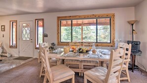 A dining area with a wooden table set for four, surrounded by wooden chairs. A large window with a wooden frame provides natural light. - Duck Creek Village Cabin w/ Deck on Half Acre (Duck Creek Village)