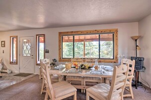 A dining area with a wooden table set for four, surrounded by wooden chairs. A large window with a wooden frame provides natural light.