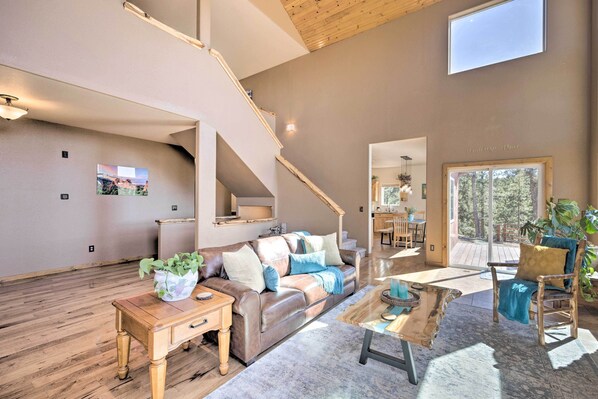 A spacious living room with a high ceiling, wooden flooring, a leather sofa, a coffee table, and a staircase leading to an upper level. - Woodland Park Home w/ Mountain Views By ATV Trails (Divide)