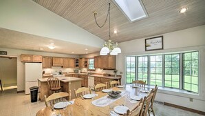 Una cocina amplia con armarios de madera, un comedor con una mesa grande preparada para seis personas, y vistas a un paisaje verde a través de ventanas grandes.