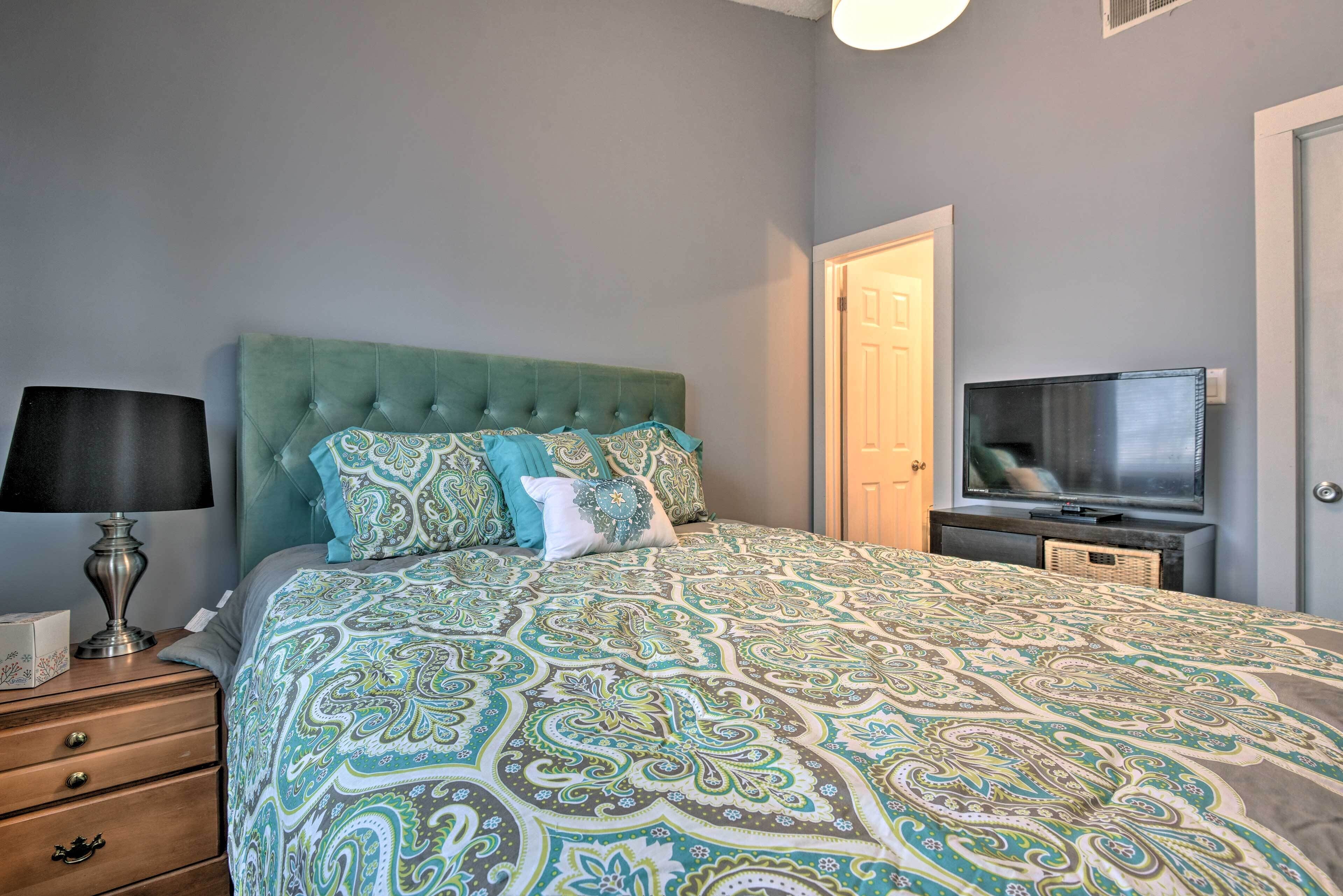 A bedroom with a tufted headboard, a patterned bedspread, a bedside lamp, a television on a stand, and a door.