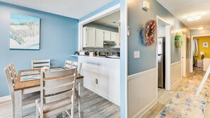 A modern kitchen with white cabinets, a wooden dining table, and a painting on the wall. - Kure Beach Condo w/ Panoramic Ocean Views! (Kure Beach)