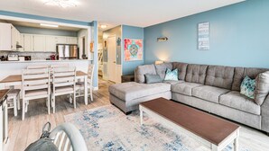 A living room with a grey sofa, a wooden coffee table, and a dining area with white chairs and a wooden table. - Kure Beach Condo w/ Panoramic Ocean Views! (Kure Beach)