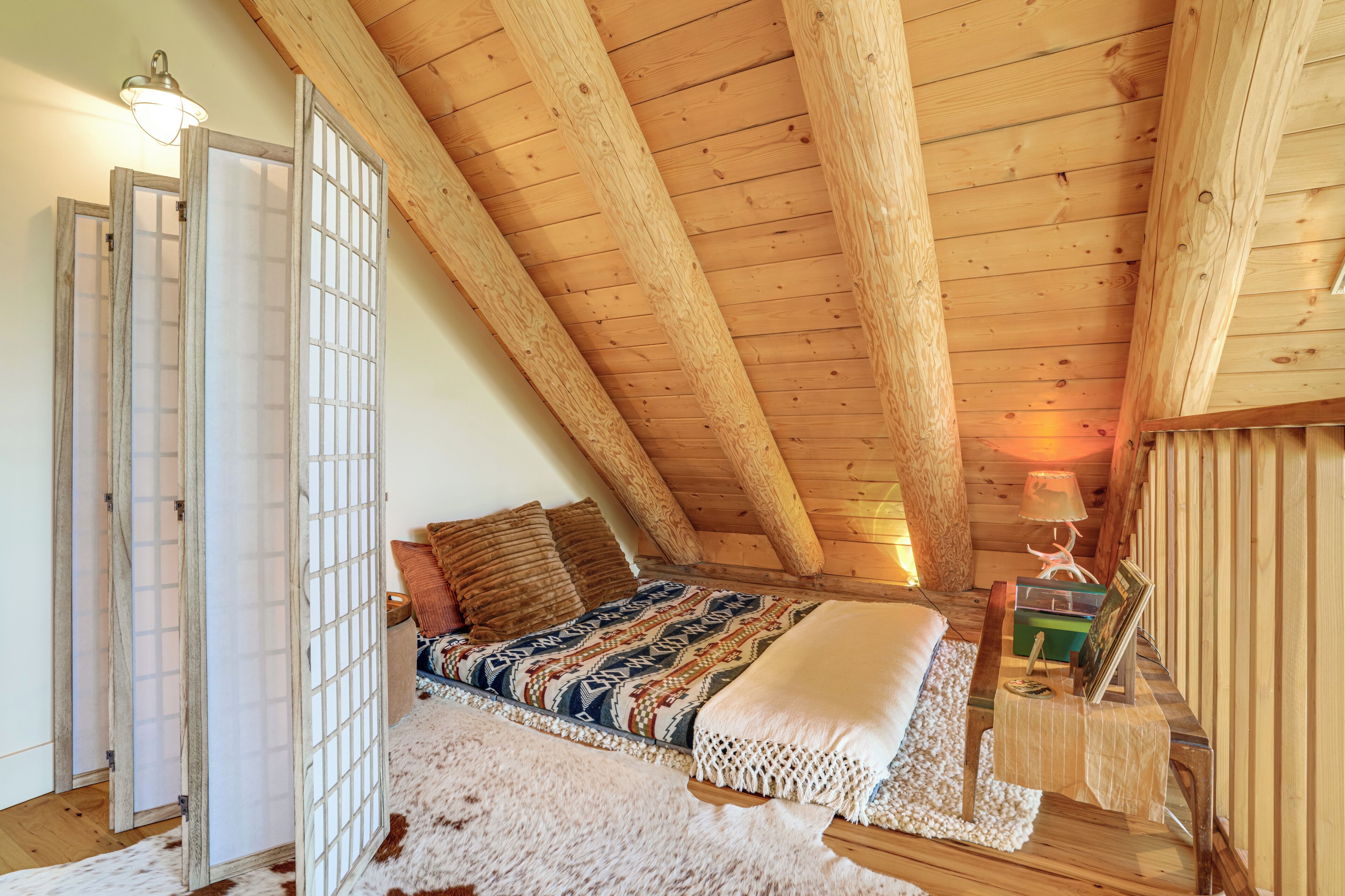 Ett mysigt sovrum på vinden med ett sluttande tak, en säng med mönstrat sänglinne, ett litet sängbord med en lampa och en dekorativ väggpanel.