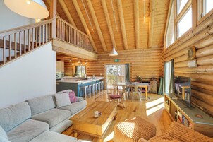 A log cabin interior with a sofa, wooden dining table, kitchen area, and a TV.