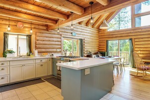 A log cabin interior with a kitchen featuring a central island, wooden beams, and large windows.