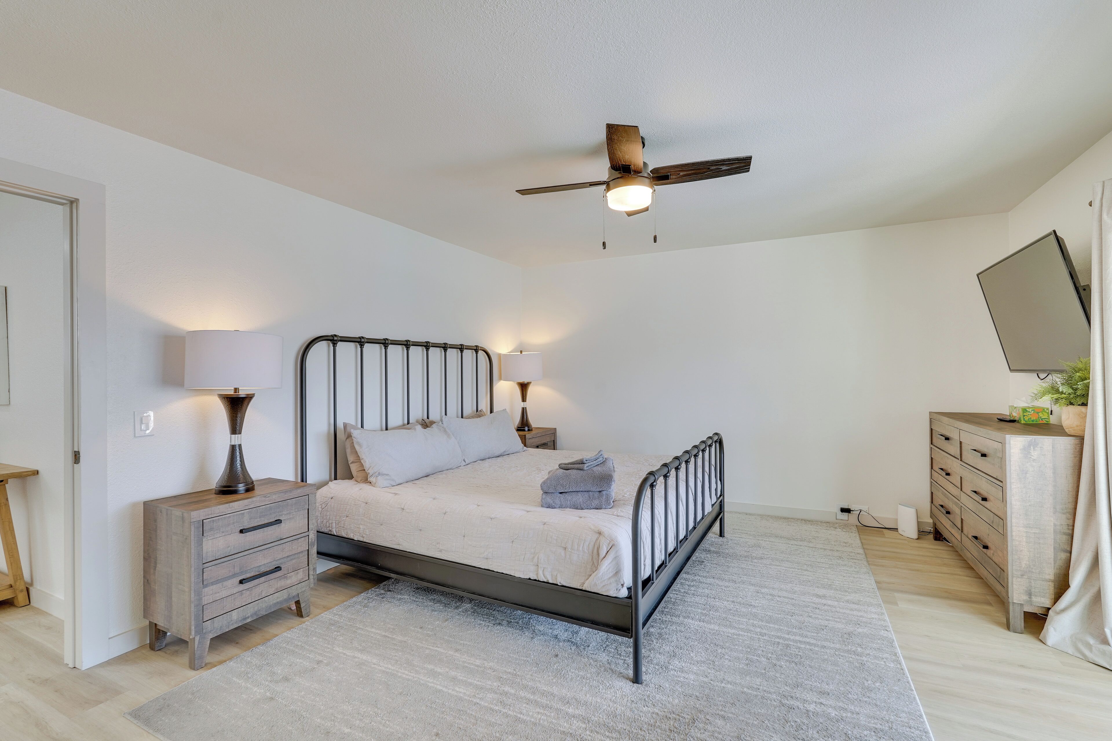 A bedroom with a bed, bedside lamps, a wooden nightstand, a dresser, a ceiling fan, and a mirror.