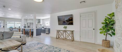 A modern living room with a sofa, coffee table, and a rug. There is a kitchen in the background with cabinets and appliances.