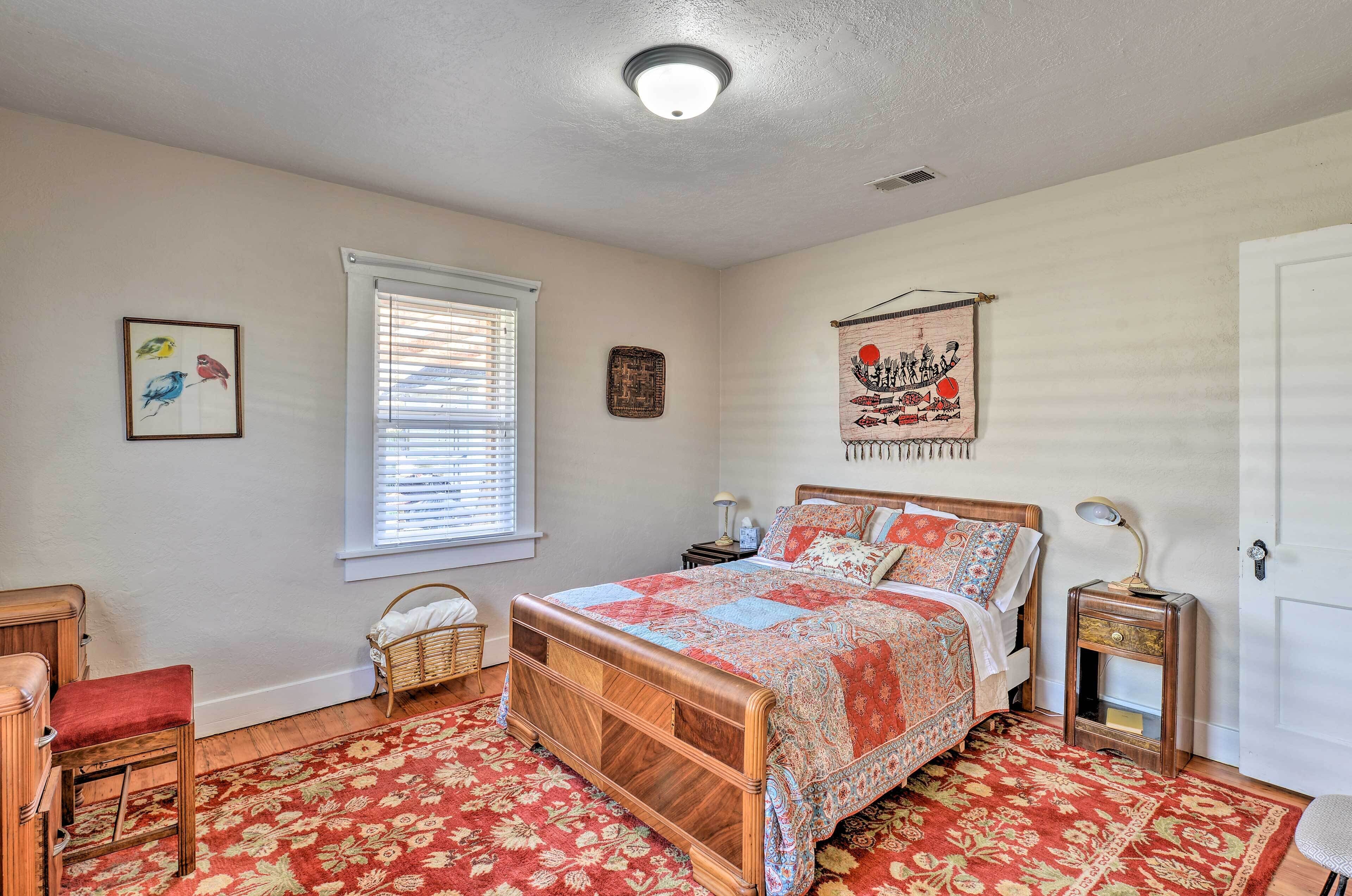 Une chambre avec un lit en bois, un tapis rouge à motifs, une fenêtre avec des stores, une décoration murale et une table de chevet avec une lampe.