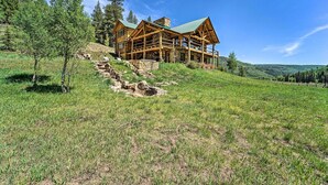 A log cabin with a stone foundation, wooden exterior, and a green roof, situated on a grassy hillside with trees around. - Fish Creek Ranch Lodge With Hot Tub and Game Room (Dolores)