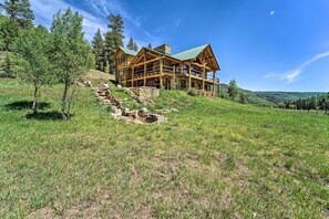 A log cabin with a stone foundation, wooden exterior, and a green roof, situated on a grassy hillside with trees around. - Fish Creek Ranch Lodge With Hot Tub and Game Room (Dolores)