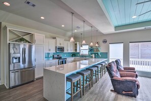 A modern kitchen with a central island, stainless steel appliances, pendant lighting, and a seating area with cushioned chairs. - Walk to Ocean: Bright Crystal Beach House w/ Deck! (Port Bolivar)