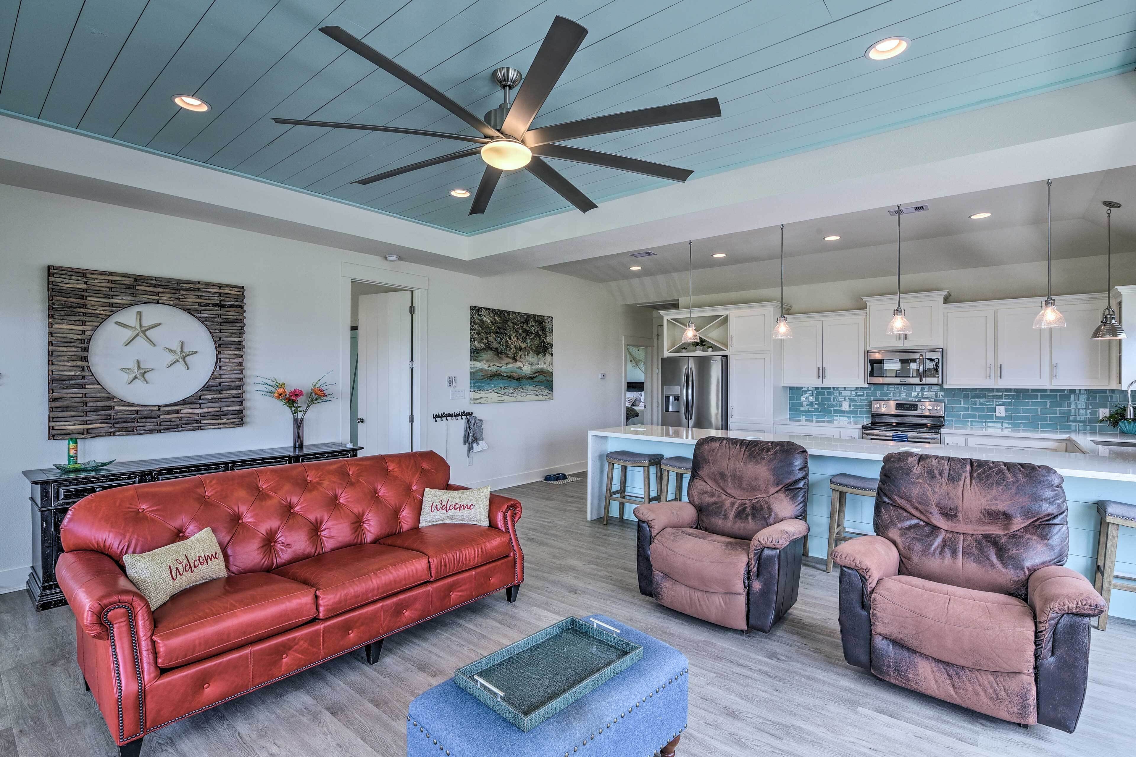 A spacious living room with a red leather sofa, two armchairs, and a ceiling fan. A kitchen with white cabinets and a blue countertop is visible in the background.