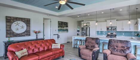 A spacious living room with a red leather sofa, two armchairs, and a ceiling fan. A kitchen with white cabinets and a blue countertop is visible in the background.