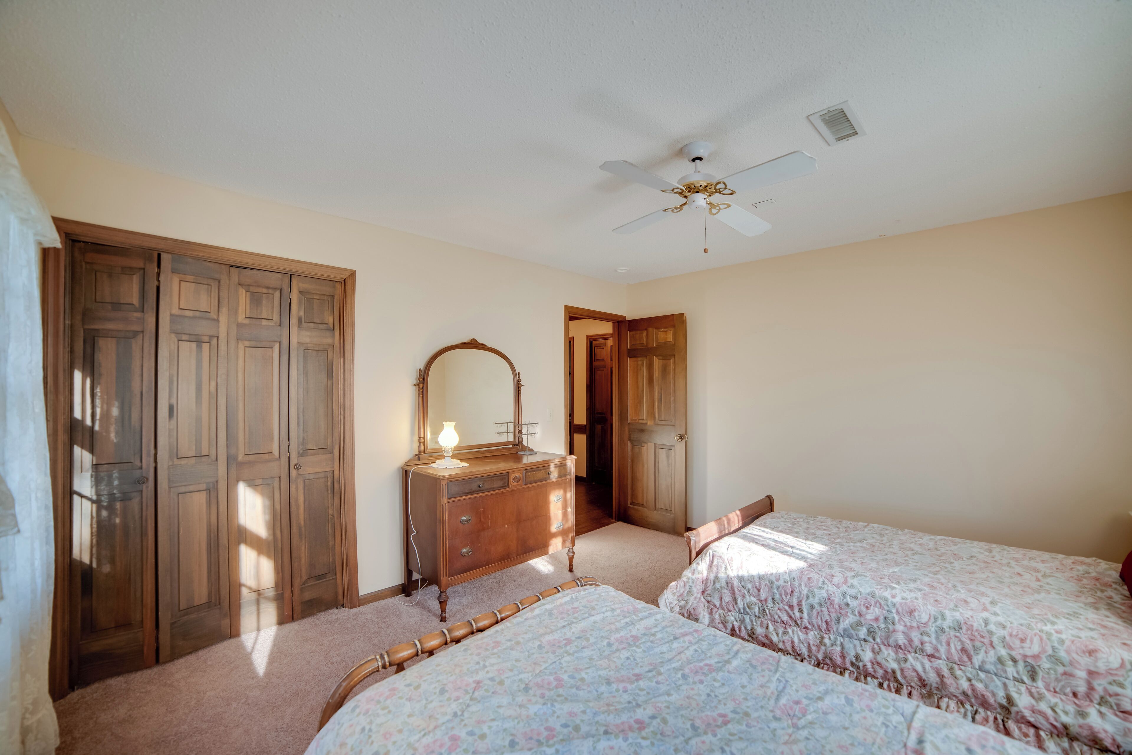 A bedroom with two beds, a wooden dresser, a ceiling fan, and a door leading to another room.
