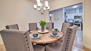 Un comedor con una mesa redonda de madera, seis sillas tapizadas, una araña y un jarrón con flores amarillas.