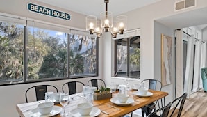 A dining area with a wooden table set for four, large windows with a view of trees, and a sign reading "BEACH HOUSE". - Walk to Coligny Beach Park: Coastal Oasis w/ Pool (Hilton Head Island)