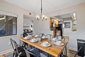 Un coin repas avec une table en bois dressée pour quatre personnes, entourée de chaises. Un lustre est suspendu au plafond, et une cuisine se trouve au fond de la pièce.