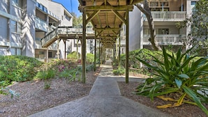 A covered walkway between buildings with a concrete path, surrounded by greenery and plants. - Walk to Coligny Beach Park: Coastal Oasis w/ Pool (Hilton Head Island)