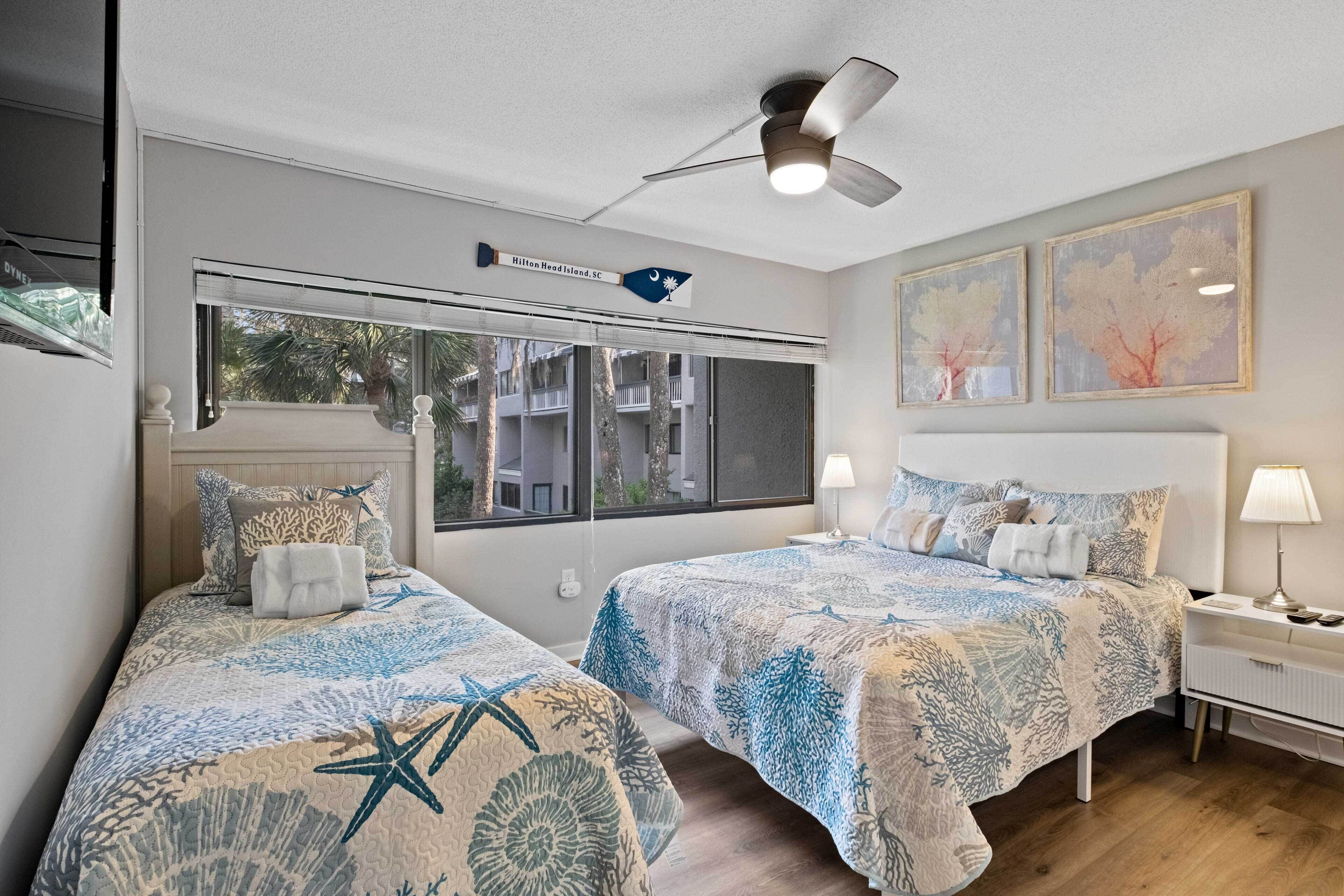 A hotel room with two beds, a ceiling fan, a window with a view of buildings, and artwork on the wall.