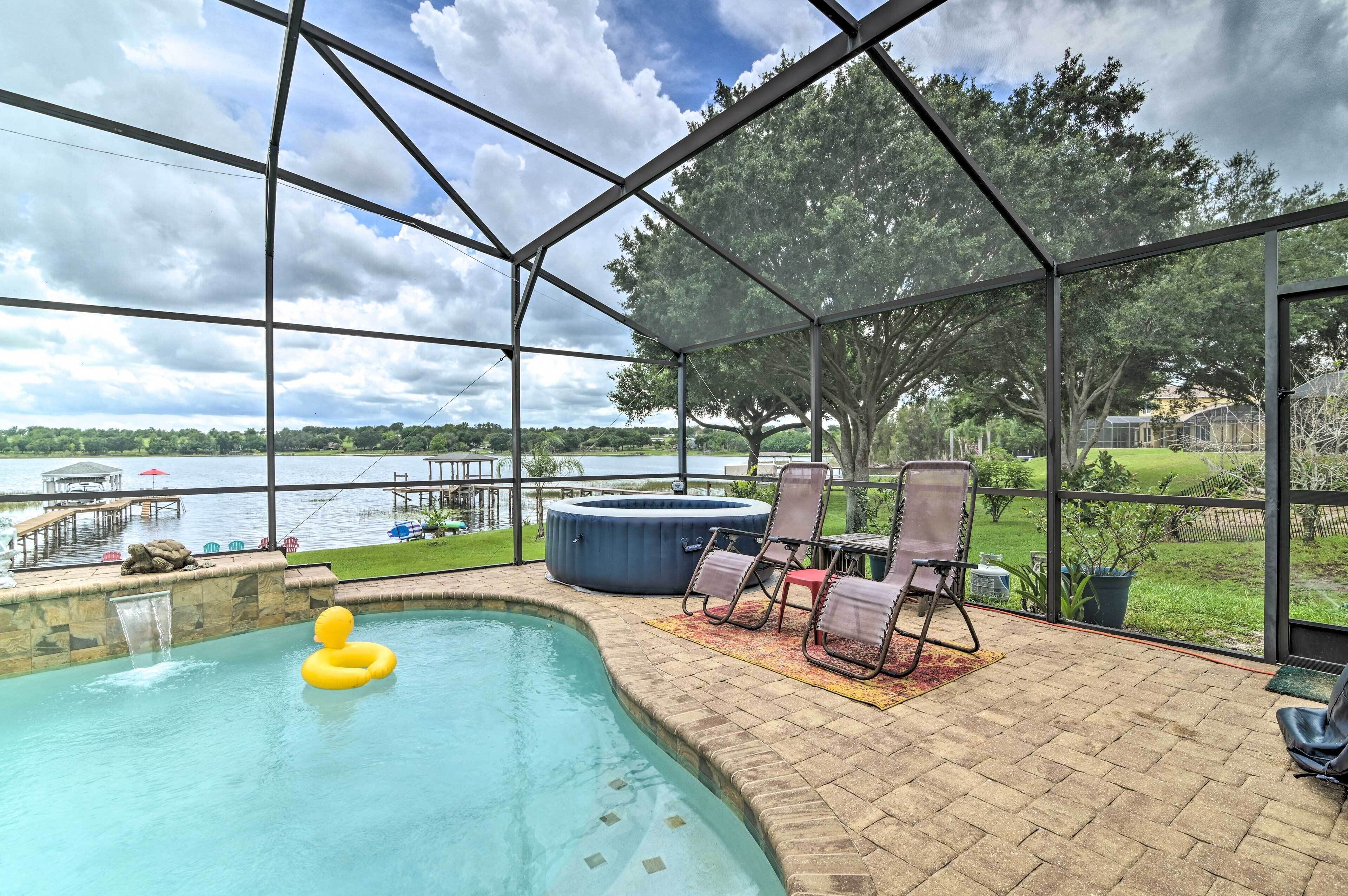 A covered outdoor swimming pool with a hot tub, lounge chairs, and a view of a lake and dock.