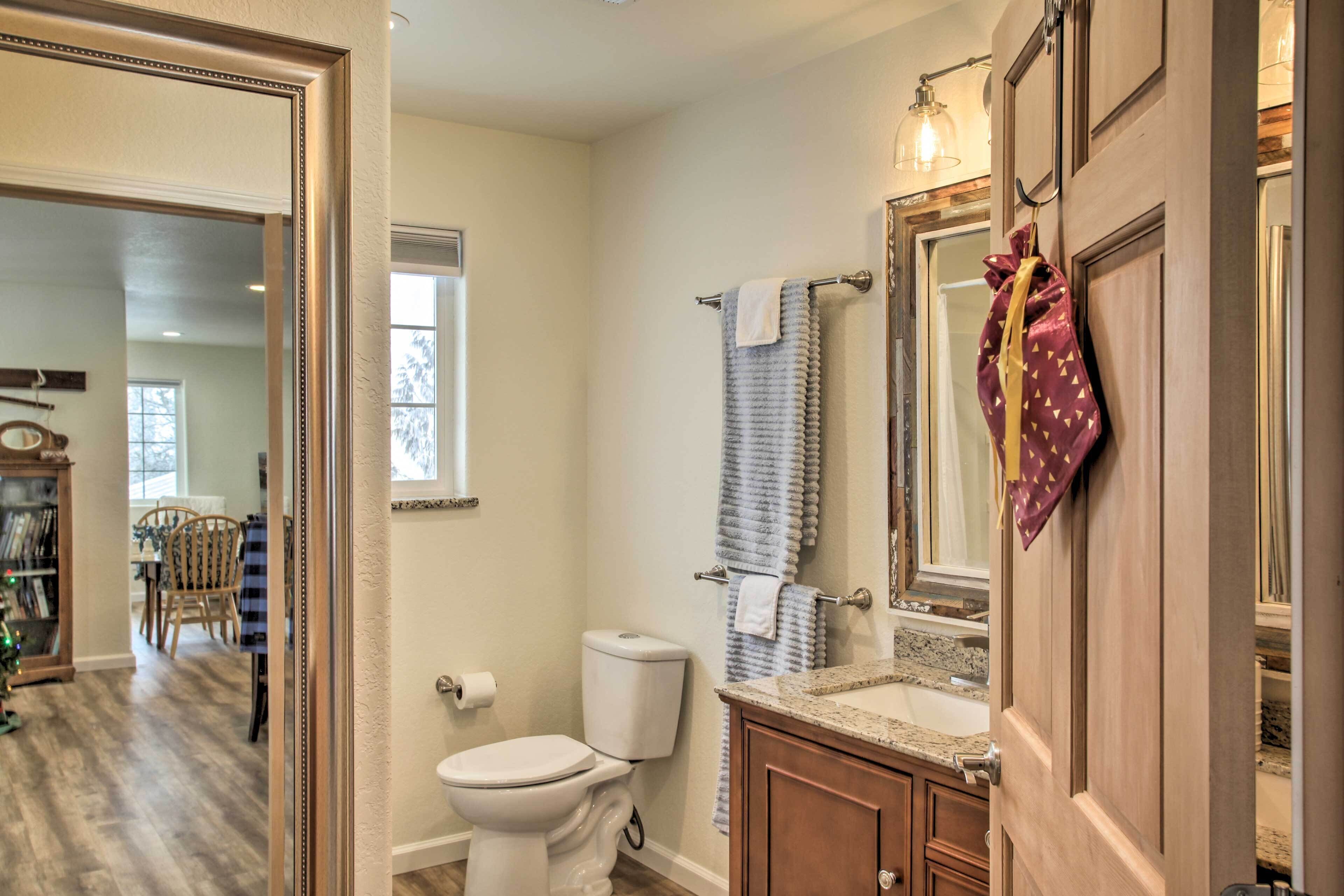 A bathroom with a toilet, sink, and mirror. There is a towel rack with towels and a bag hanging on it. The bathroom has wooden cabinets and a window.