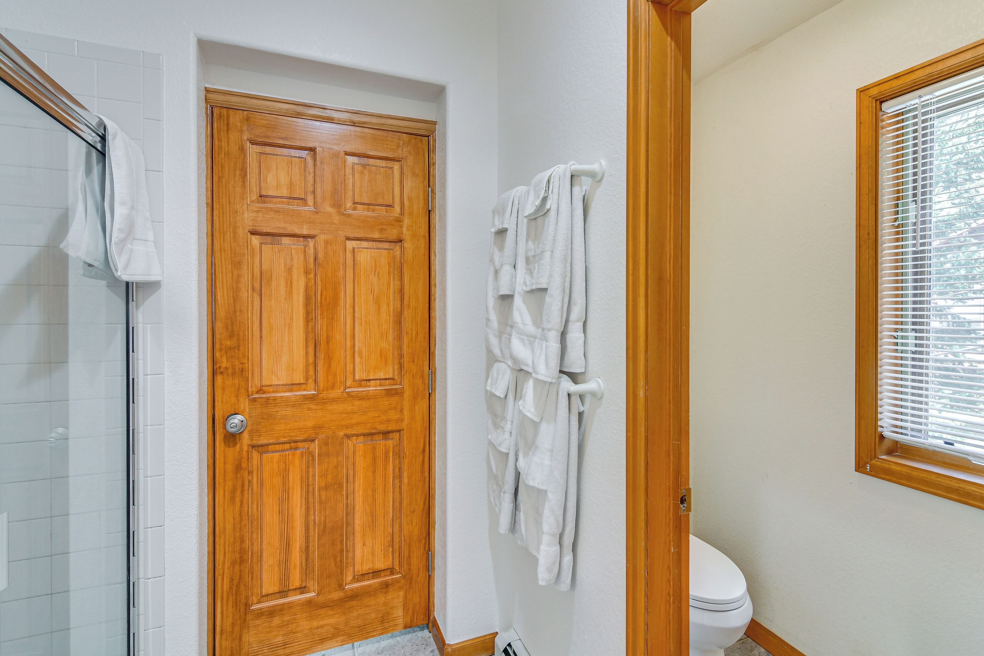 A bathroom with a wooden door, a towel rack with multiple towels, a toilet, and a window with blinds.
