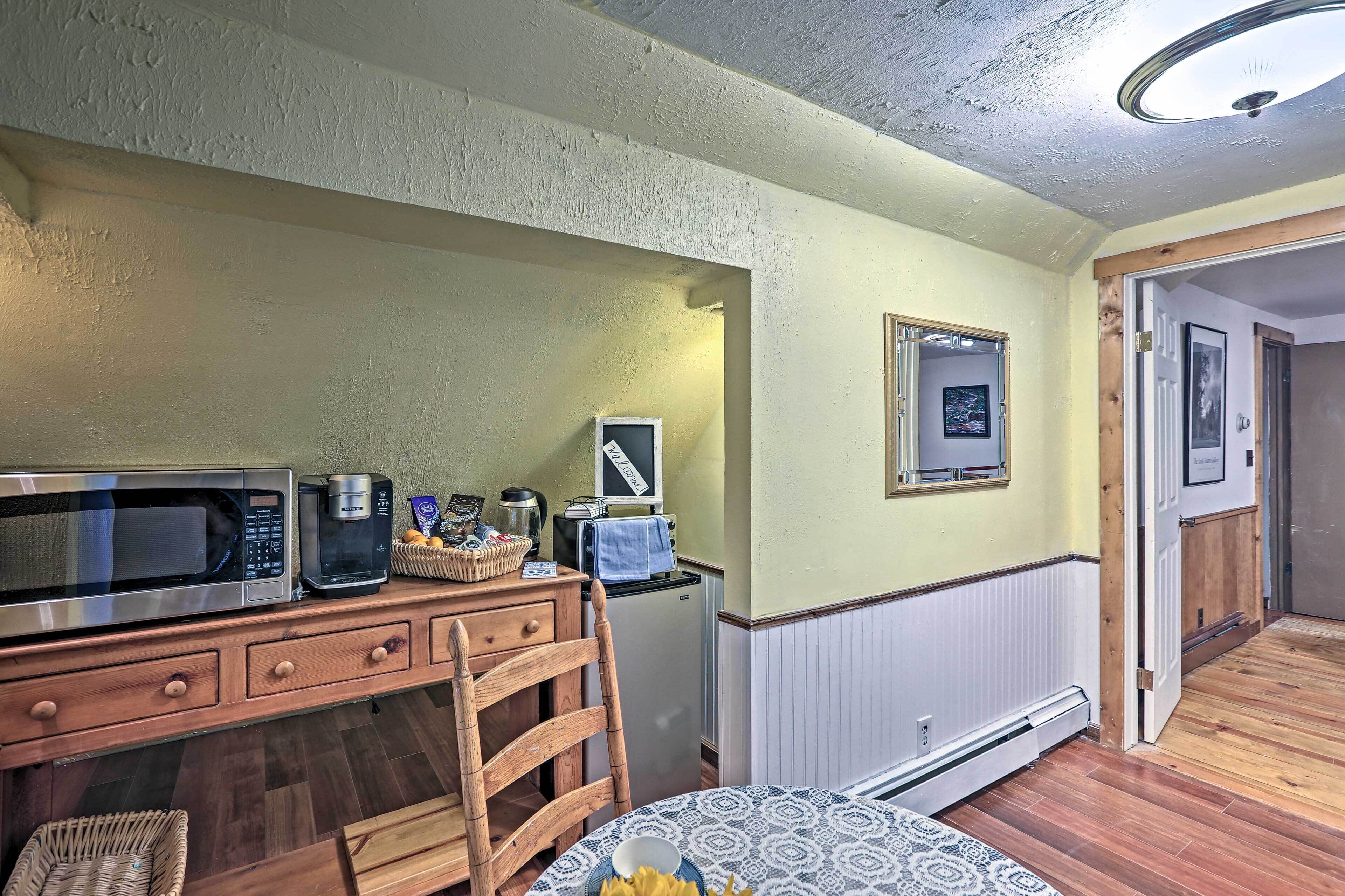 Eine kleine Küche mit Mikrowelle, Kaffeemaschine und Toaster auf einer Holzausziehtischplatte. Ein Stuhl und ein kleiner Tisch mit gemustertem Tischläufer sind ebenfalls zu sehen.
