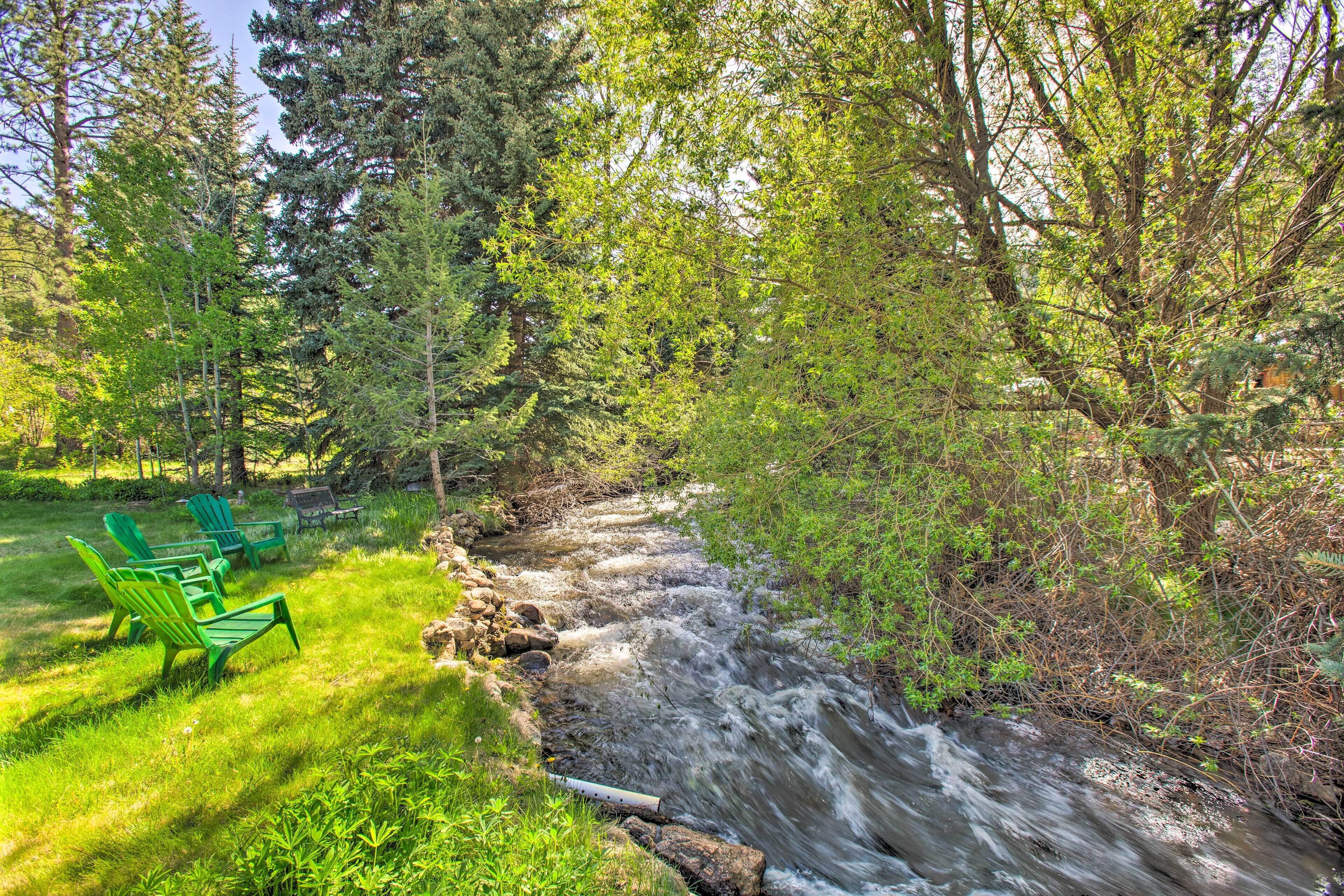 A river flowing through a forest with green chairs and a wooden bench along the riverbank.