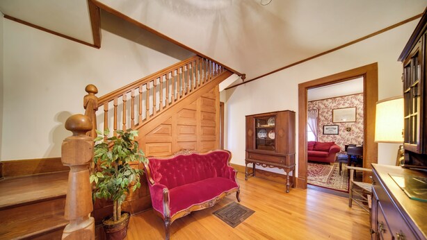 A spacious living room with a wooden staircase, a red velvet sofa, a potted plant, and a wooden cabinet. - Historic Victorian Farmhouse w/ Porch & Views! (Mayville)