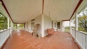 A covered porch with a swing chair and a wicker chair, white railings, and a view of trees. - Historic Victorian Farmhouse w/ Porch & Views! (Mayville)
