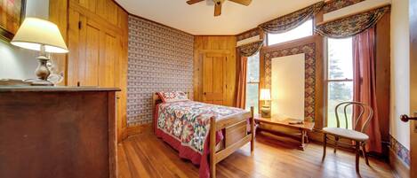 A bedroom with wooden walls, a bed with a floral bedspread, a chair, a desk with a lamp, and a window with curtains.
