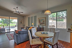 A modern living room with a grey sofa, a round dining table with chairs, a ceiling fan, and large windows with a view of the outdoors. - Zen Phoenix Home on Legacy Golf Course w/ Pool! (Phoenix)