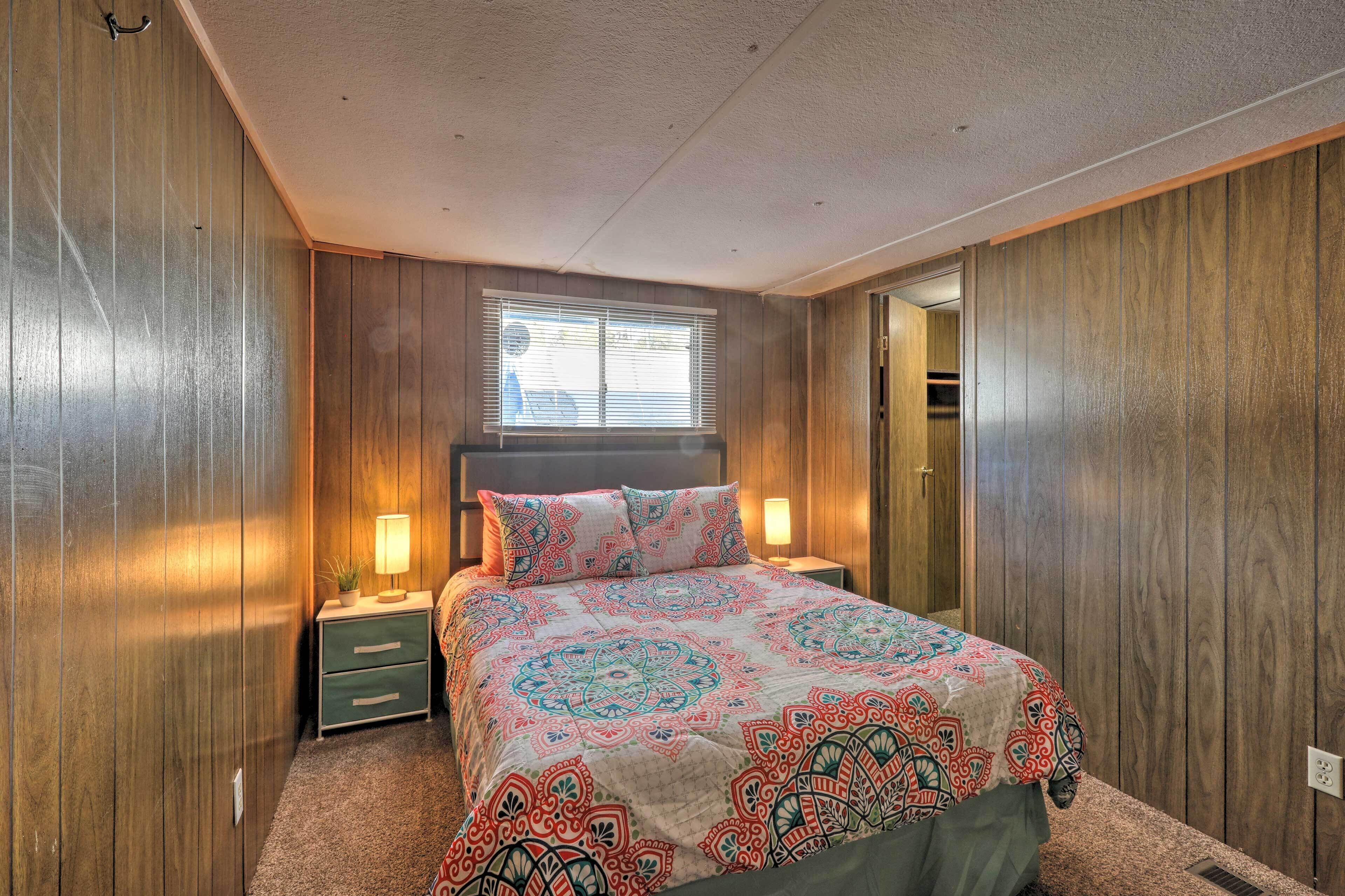 Un dormitorio con pared de madera empapelada, una cama con colcha estampada, una mesita de noche con lámpara y una ventana con persianas.