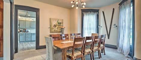 A dining room with a wooden table and chairs, a chandelier, and a view of the kitchen through an open door.