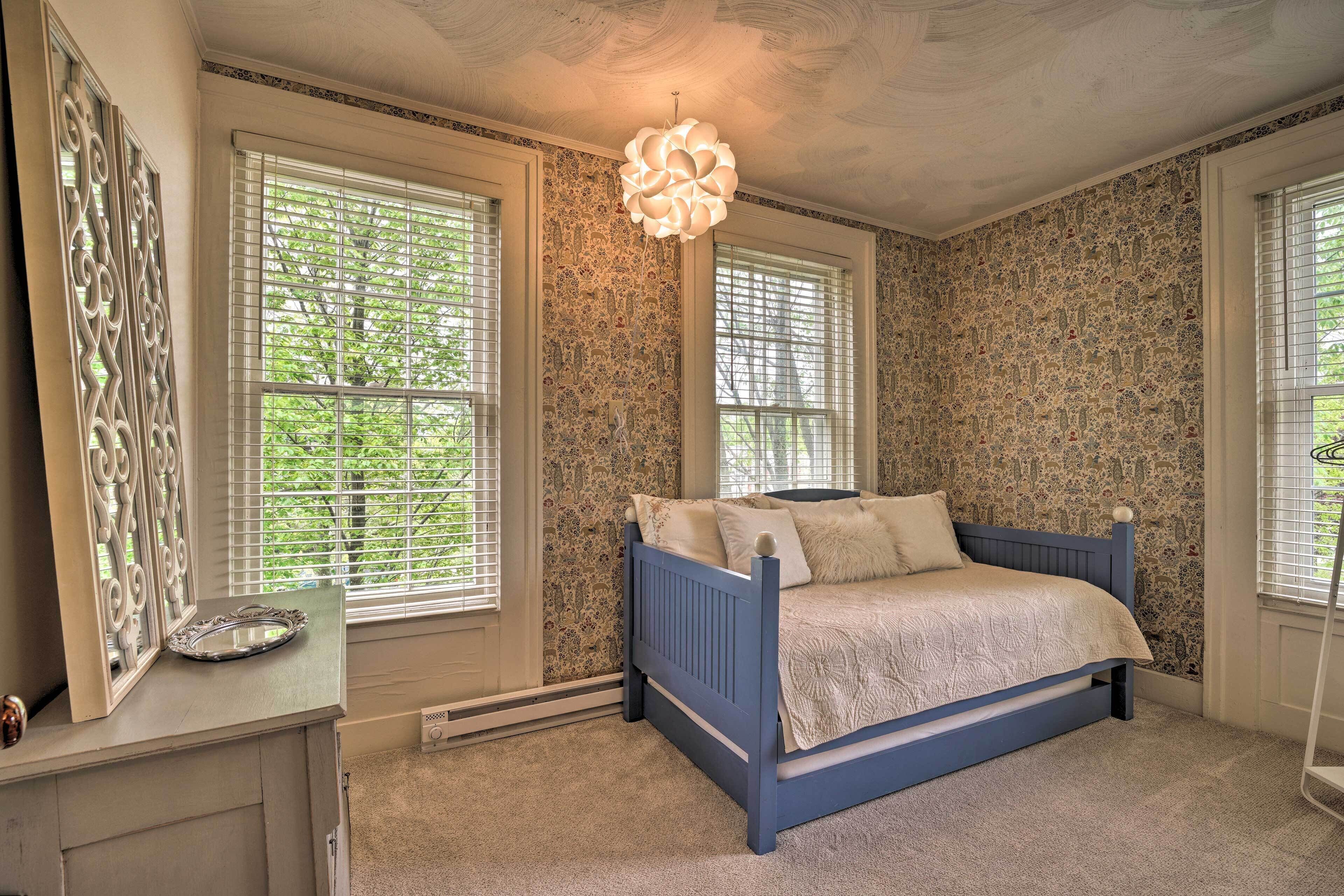 A bedroom with a blue bed, large windows with blinds, a decorative mirror, and a unique ceiling light fixture.