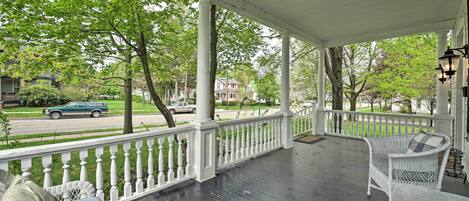 A covered porch with white railings, wicker furniture, and a view of a street with parked cars and houses.
