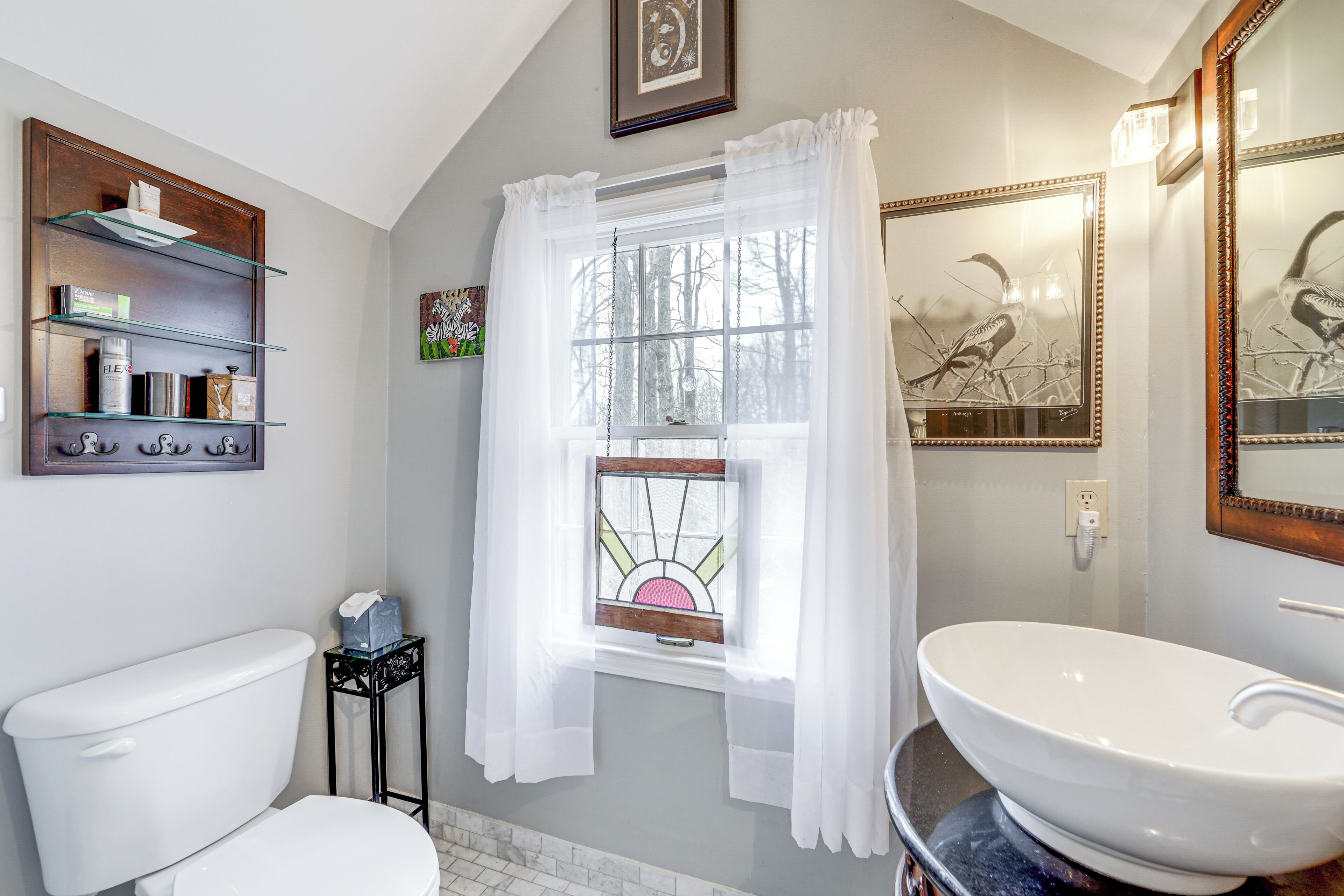 A bathroom with a white toilet, a white sink, a window with a stained glass view, and a shelf with toiletries.