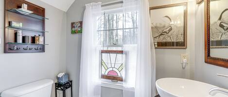 A bathroom with a white toilet, a white sink, a window with a stained glass view, and a shelf with toiletries.
