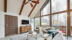 A spacious living room with a large window, a ceiling fan, a television, and a cozy seating area. - Breathtaking Brevard Home w/ Screened Porch! (Brevard)