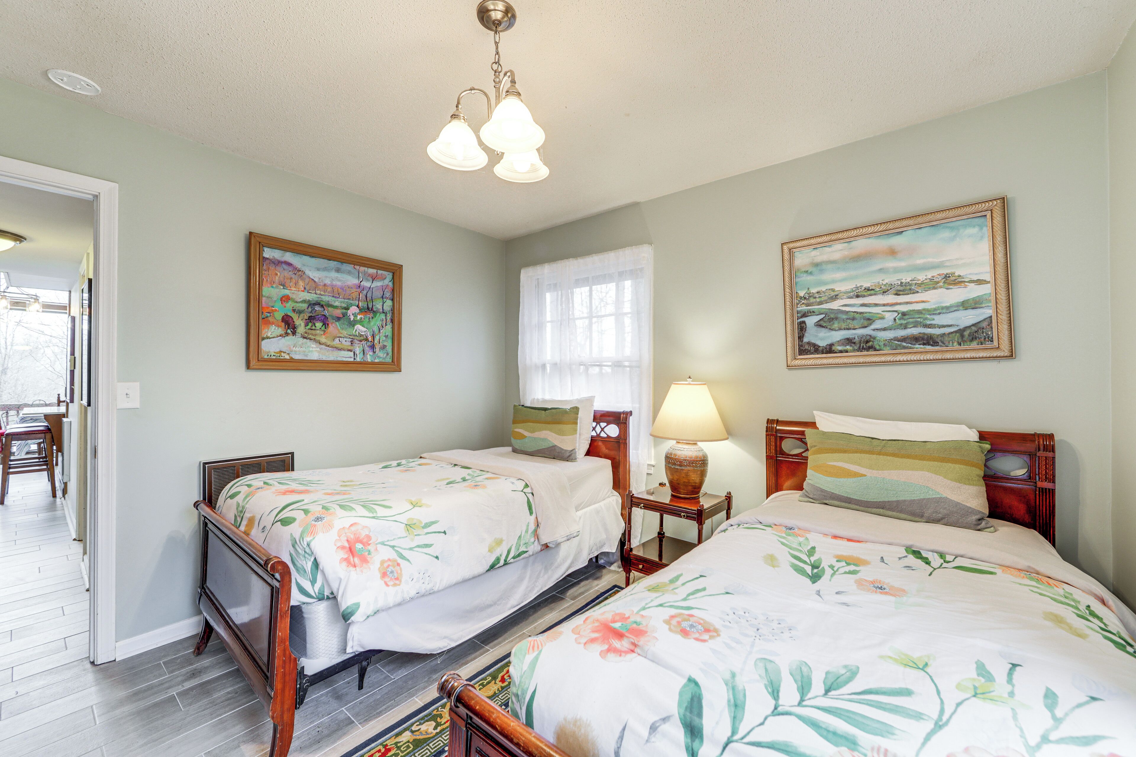 A bedroom with two wooden beds, floral bedding, a nightstand with a lamp, and two wall-mounted paintings.