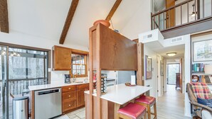 A modern kitchen with wooden cabinets, a white countertop, and a built-in dishwasher. There are two stools with red cushions and a wooden rocking chair with a plaid blanket. - Breathtaking Brevard Home w/ Screened Porch! (Brevard)