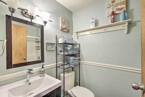 A bathroom with a white sink, a toilet, a mirror with lights, a shelf with toiletries, and a towel rack. - Bright Pine Cabin: Direct Trail Access & Deck! (Pine)