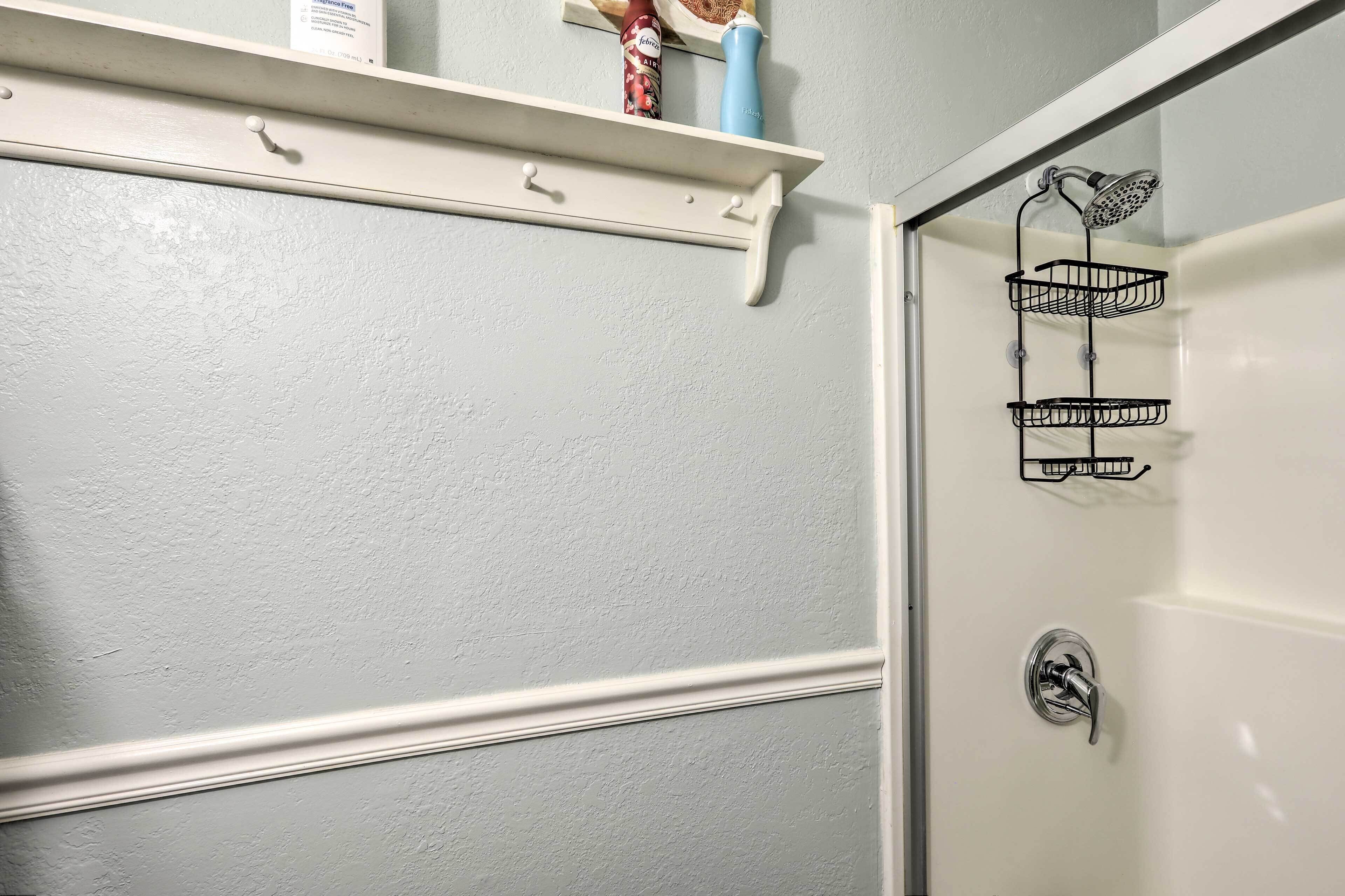 Un baño con pared blanca, zona de ducha y estante con artículos de aseo.