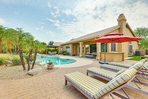 Un espace aménagé au bord de la piscine, avec des chaises longues, un parasol rouge et une maison dont le toit est recouvert de tuiles.