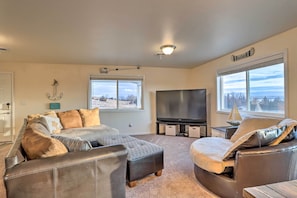 A living room with a large flat-screen TV, a sofa, an ottoman, and a chair. There are two windows providing a view of the outdoors.