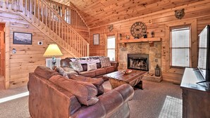 L’intérieur d’un chalet en bois comprend un escalier, une cheminée en pierre, un canapé en cuir marron et une table basse en bois.