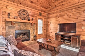A cozy log cabin interior with a stone fireplace, a large wooden dining table, a comfortable sofa, and a flat-screen TV mounted on the wall. - Sevierville Cabin w/ Lake Access & Private Hot Tub (Sevierville)